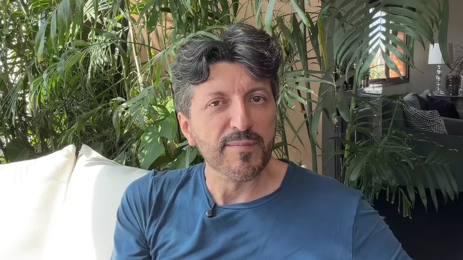 Homem de meia-idade com barba grisalha e camiseta azul em ambiente com plantas e janela