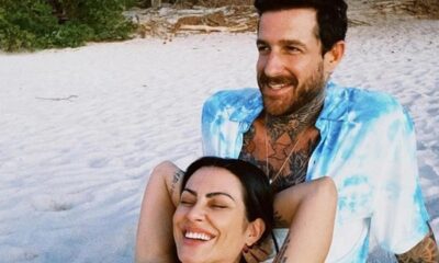 Casal sorridente na praia, mulher deitada no colo do homem tatuado, areia e vegetação ao fundo