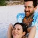 Casal sorridente na praia, mulher deitada no colo do homem tatuado, areia e vegetação ao fundo
