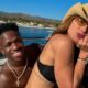 Homem sorridente e mulher de chapéu e óculos posando em barco com mar e montanhas ao fundo
