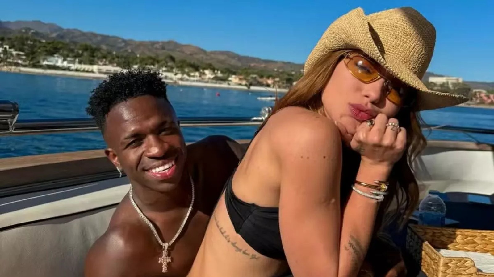 Homem sorridente e mulher de chapéu e óculos posando em barco com mar e montanhas ao fundo