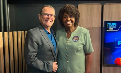 Duas pessoas sorrindo em estúdio, mulher com broche 'BENEDITA', ao lado de tela Tupi Eleições 2026