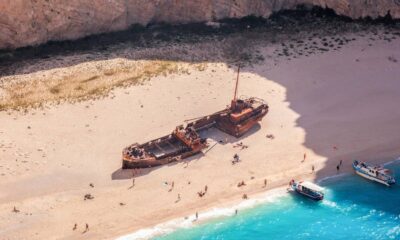 Praia com navio naufragado, pessoas na areia, barcos em água azul-turquesa, falésia rochosa ao fundo