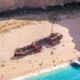 Praia com navio naufragado, pessoas na areia, barcos em água azul-turquesa, falésia rochosa ao fundo