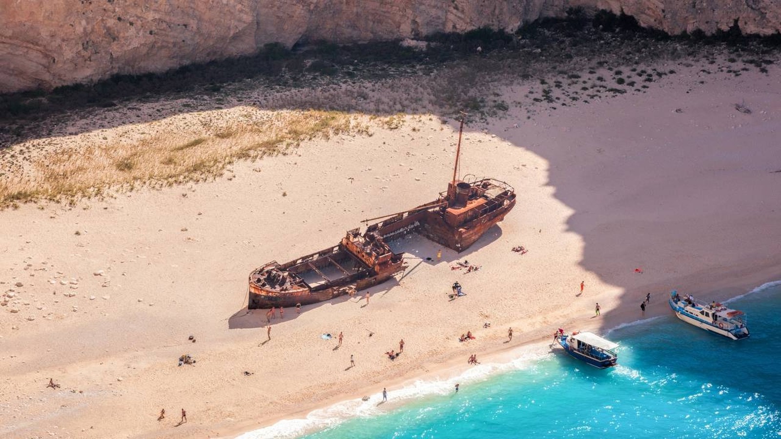Praia com navio naufragado, pessoas na areia, barcos em água azul-turquesa, falésia rochosa ao fundo