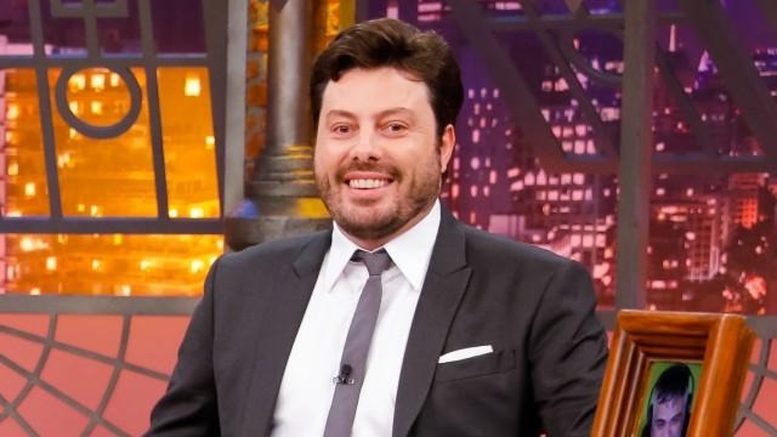 Danilo Gentili, de terno e gravata, sorri para a câmera em cenário de televisão