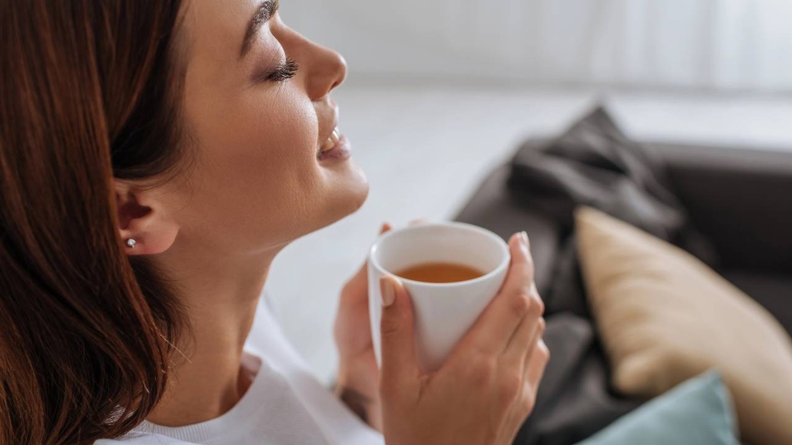Mulher sorrindo com olhos fechados, segurando uma xícara de chá quente em ambiente doméstico
