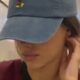 Bruna Biancardi de boné azul e camiseta vinho cobrindo o rosto, mão na orelha