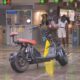 Scooter elétrica preta com assentos marrons estacionada em terminal de barcas, catracas e pessoas ao fundo
