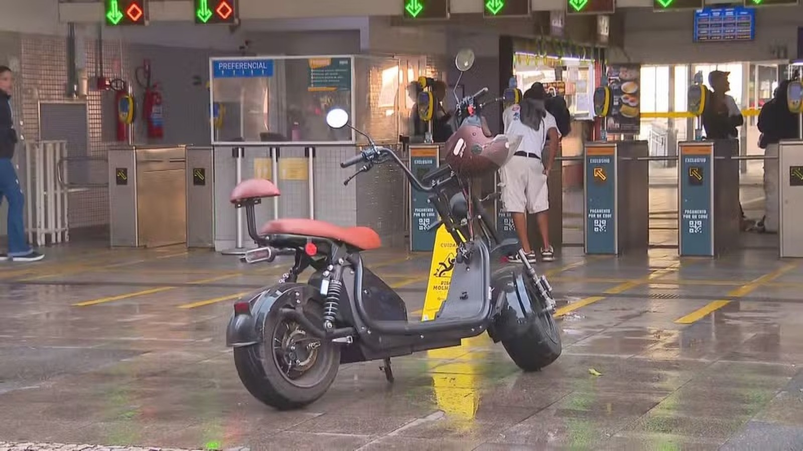 Scooter elétrica preta com assentos marrons estacionada em terminal de barcas, catracas e pessoas ao fundo