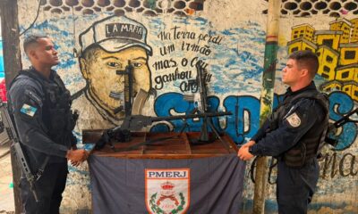 Dois policiais do PMERJ exibem fuzis e uma metralhadora apreendidos em frente a um muro grafitado