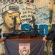 Dois policiais do PMERJ exibem fuzis e uma metralhadora apreendidos em frente a um muro grafitado