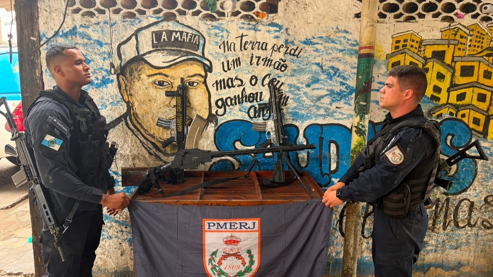 Dois policiais do PMERJ exibem fuzis e uma metralhadora apreendidos em frente a um muro grafitado