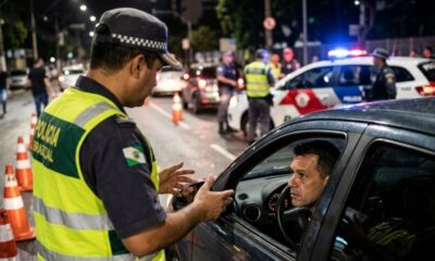 Policial com colete amarelo aborda motorista em carro à noite durante blitz