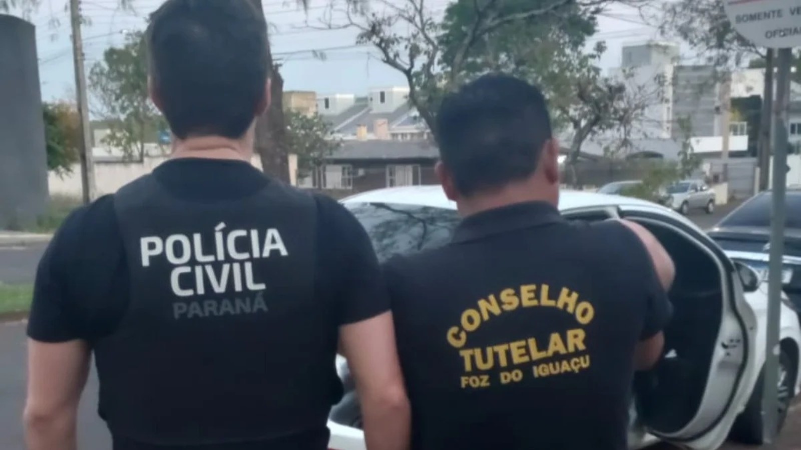 Homens da Polícia Civil e Conselho Tutelar, de costas, perto de carro branco em rua