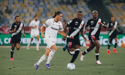 Jogador Fluminense camisa branca com bola e três Vasco camisa preta em campo de futebol