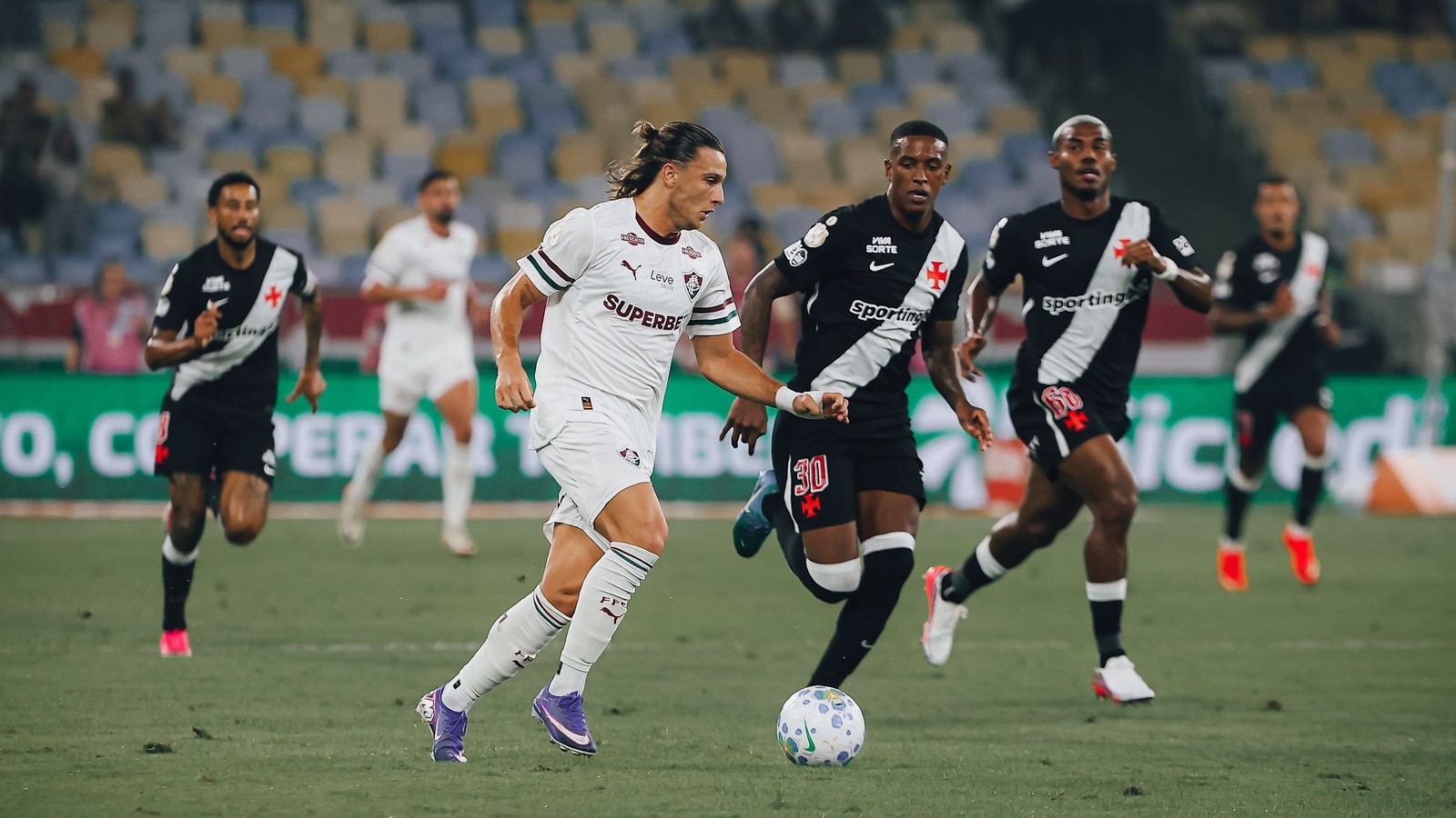 Jogador Fluminense camisa branca com bola e três Vasco camisa preta em campo de futebol