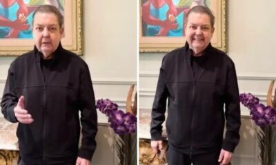 Faustão sorri e gesticula em vídeo, vestindo jaqueta preta, com quadro e flores ao fundo