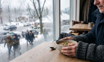 Pessoa segura tigela de sopa em mesa de madeira, janela com gotas de chuva e neve lá fora