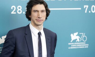Adam Driver de terno azul, camisa branca, gravata, sorrindo em fundo azul com ‘28. 7.09’