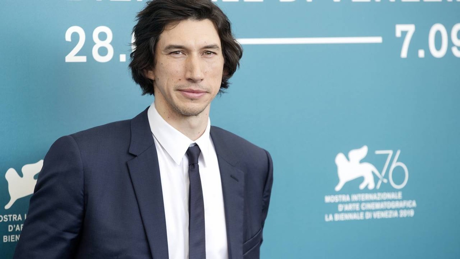 Adam Driver de terno azul, camisa branca, gravata, sorrindo em fundo azul com ‘28. 7.09’