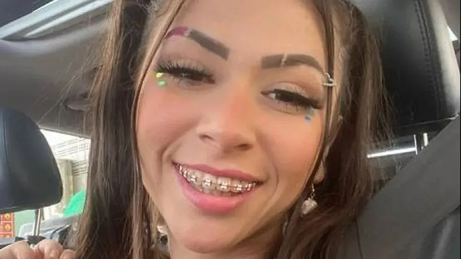 Jovem mulher sorrindo com cabelos longos, aparelho ortodôntico, maquiagem vibrante e piercing na sobrancelha