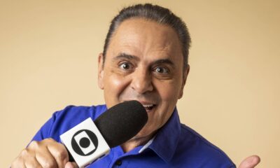 Luis Roberto sorrindo, olhos arregalados, segurando microfone com logo da Globo e fundo bege