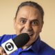 Luis Roberto sorrindo, olhos arregalados, segurando microfone com logo da Globo e fundo bege