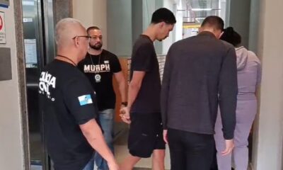 Policiais civis, um com camiseta POLÍCIA CIVIL, escoltam três pessoas, uma delas algemada