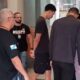 Policiais civis, um com camiseta POLÍCIA CIVIL, escoltam três pessoas, uma delas algemada