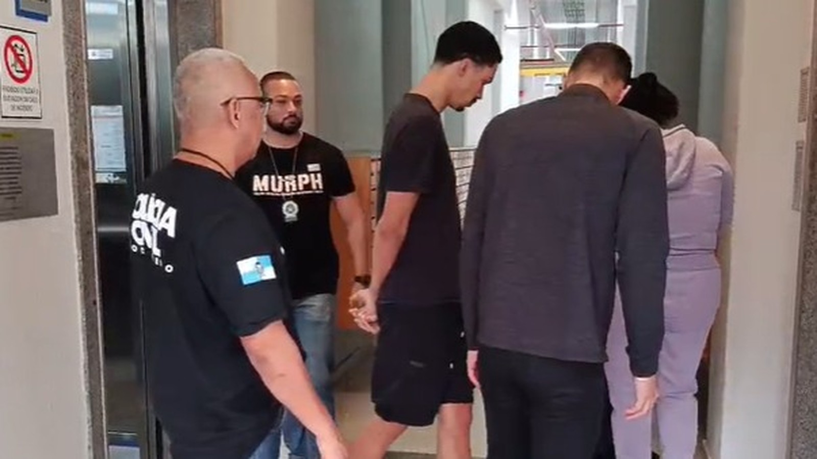Policiais civis, um com camiseta POLÍCIA CIVIL, escoltam três pessoas, uma delas algemada