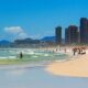 Praia movimentada com mar agitado, areia, pessoas, guarda-sóis coloridos e prédios altos