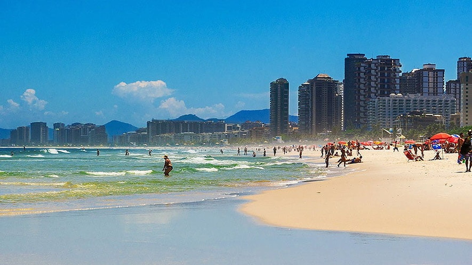 Praia movimentada com mar agitado, areia, pessoas, guarda-sóis coloridos e prédios altos