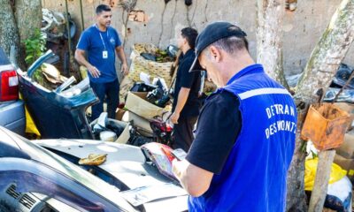 Agente do Detran RJ de costas com colete azul inspeciona peças de carro em ferro-velho