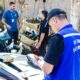 Agente do Detran RJ de costas com colete azul inspeciona peças de carro em ferro-velho