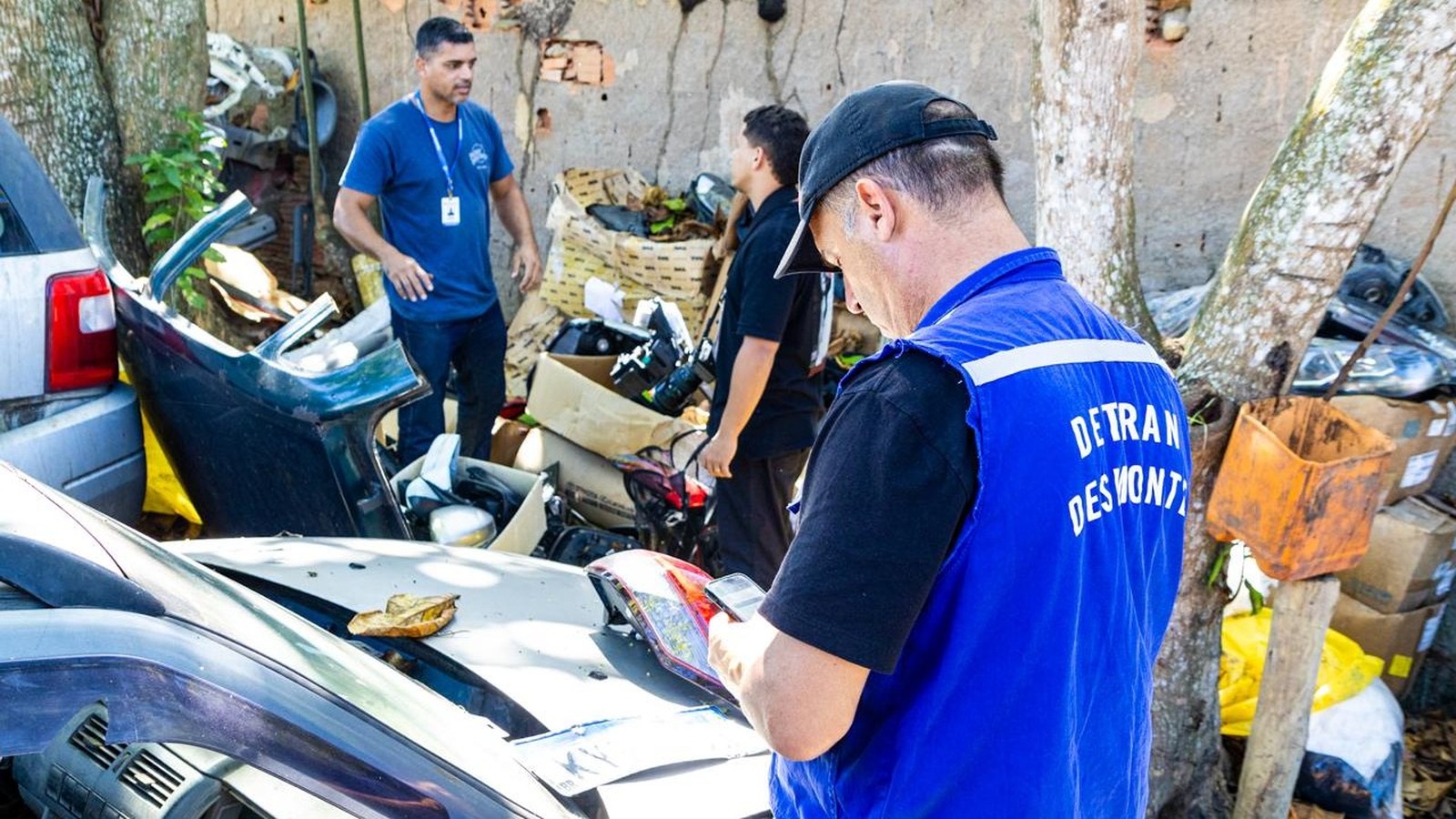 Agente do Detran RJ de costas com colete azul inspeciona peças de carro em ferro-velho