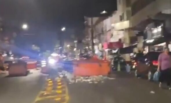 Rua noturna com blocos grandes laranja, detritos, motocicleta, carro, pessoa e edifícios