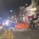 Rua noturna com blocos grandes laranja, detritos, motocicleta, carro, pessoa e edifícios