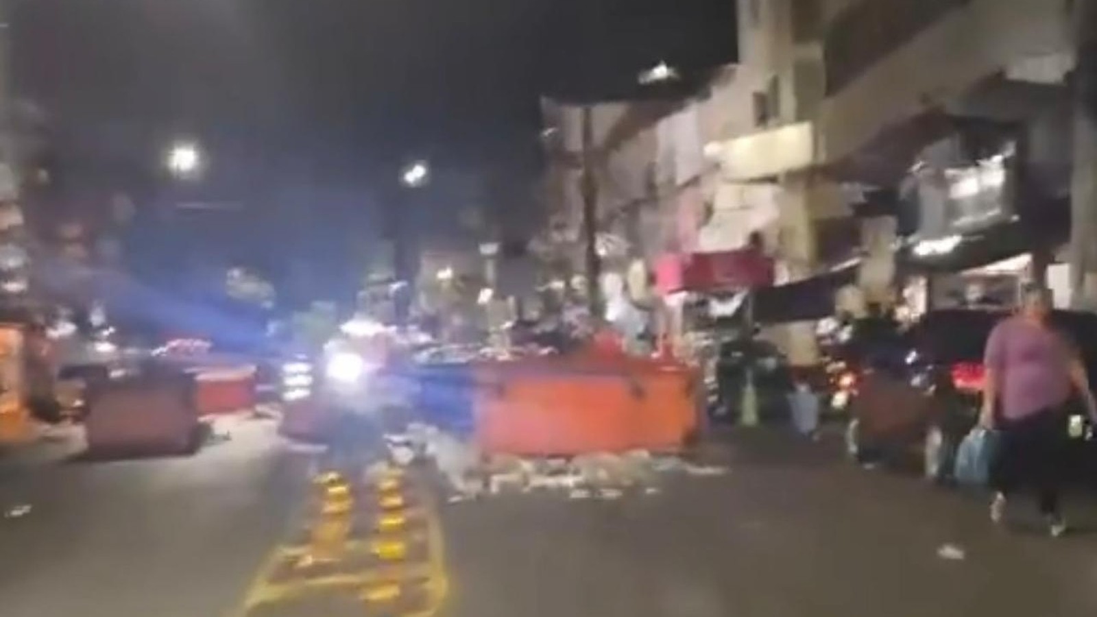 Rua noturna com blocos grandes laranja, detritos, motocicleta, carro, pessoa e edifícios