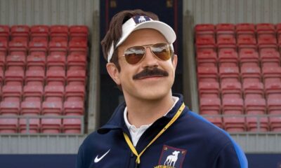 Homem sorridente com bigode, óculos de sol e boné, vestindo agasalho azul em estádio