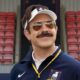 Homem sorridente com bigode, óculos de sol e boné, vestindo agasalho azul em estádio