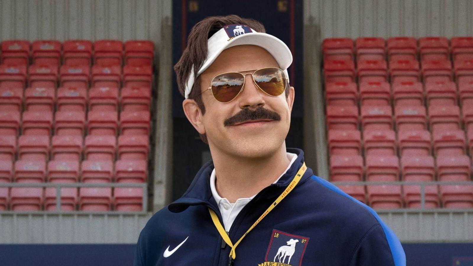 Homem sorridente com bigode, óculos de sol e boné, vestindo agasalho azul em estádio