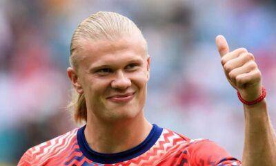 Close-up de Erling Haaland sorrindo, cabelo loiro preso, fazendo sinal de positivo com a mão direita