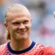 Close-up de Erling Haaland sorrindo, cabelo loiro preso, fazendo sinal de positivo com a mão direita