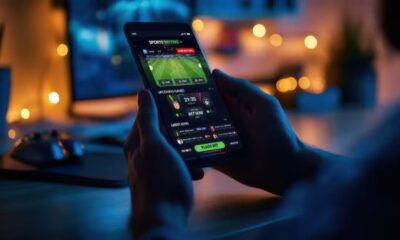 Mãos seguram smartphone exibindo aplicativo de apostas esportivas em ambiente escuro com luzes