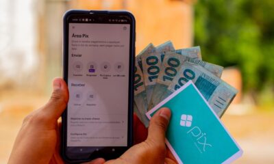 Mãos seguram celular com app Pix aberto e notas de R$100 com cartão Pix
