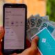 Mãos seguram celular com app Pix aberto e notas de R$100 com cartão Pix