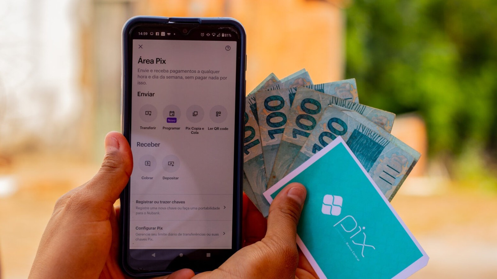 Mãos seguram celular com app Pix aberto e notas de R$100 com cartão Pix