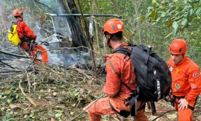 Resgatistas em floresta, com fumaça e destroços de helicóptero queimado ao fundo