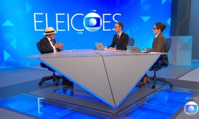 Lula, com chapéu, e dois jornalistas em estúdio de TV, com 'ELEICOES' ao fundo